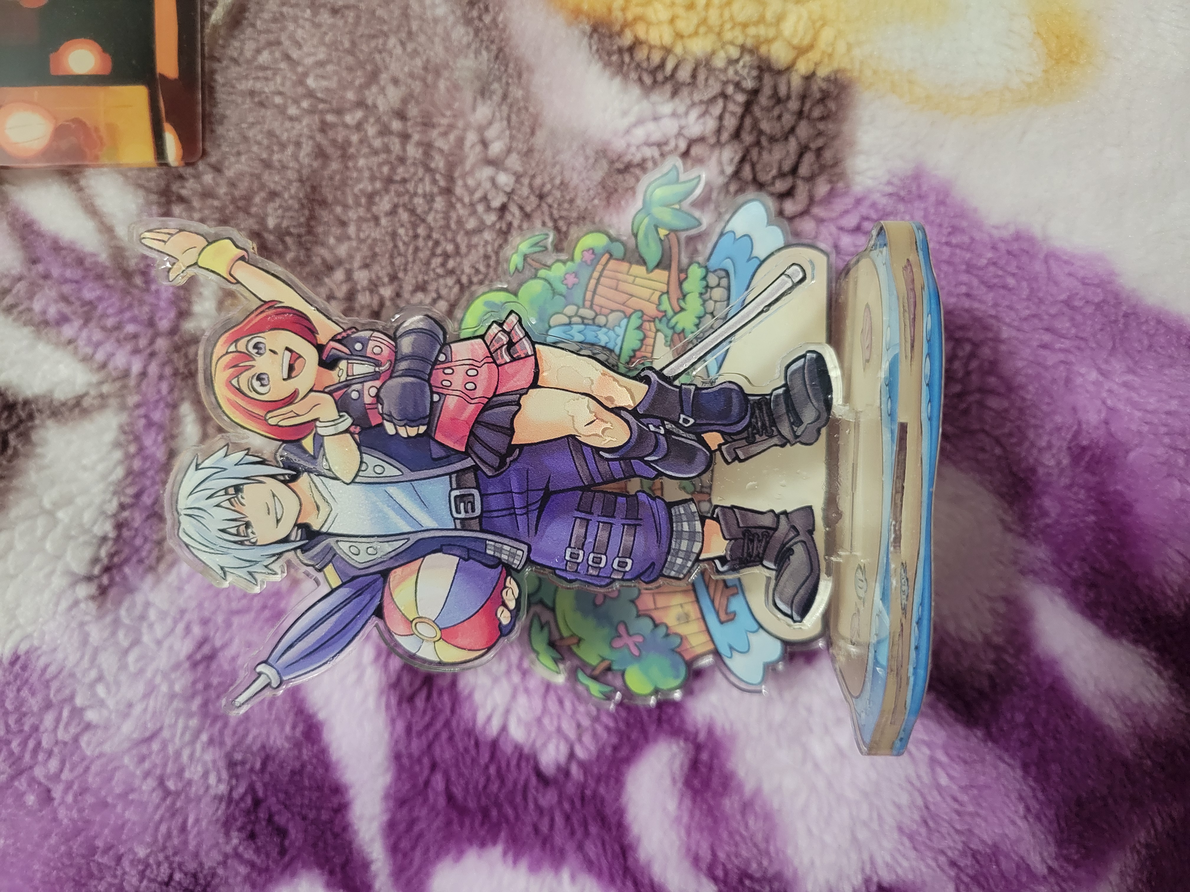 Two piece acrylic standee of Riku holding a beach ball and umbrella in one hand and Kairi by her waist in the other. Kairi shouts with her arm in a wave while Destiny Island is in the background and they stand on the shoreline.